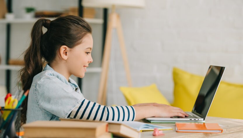 side view of adorable child using laptop while sitting at desk with books and doing homework